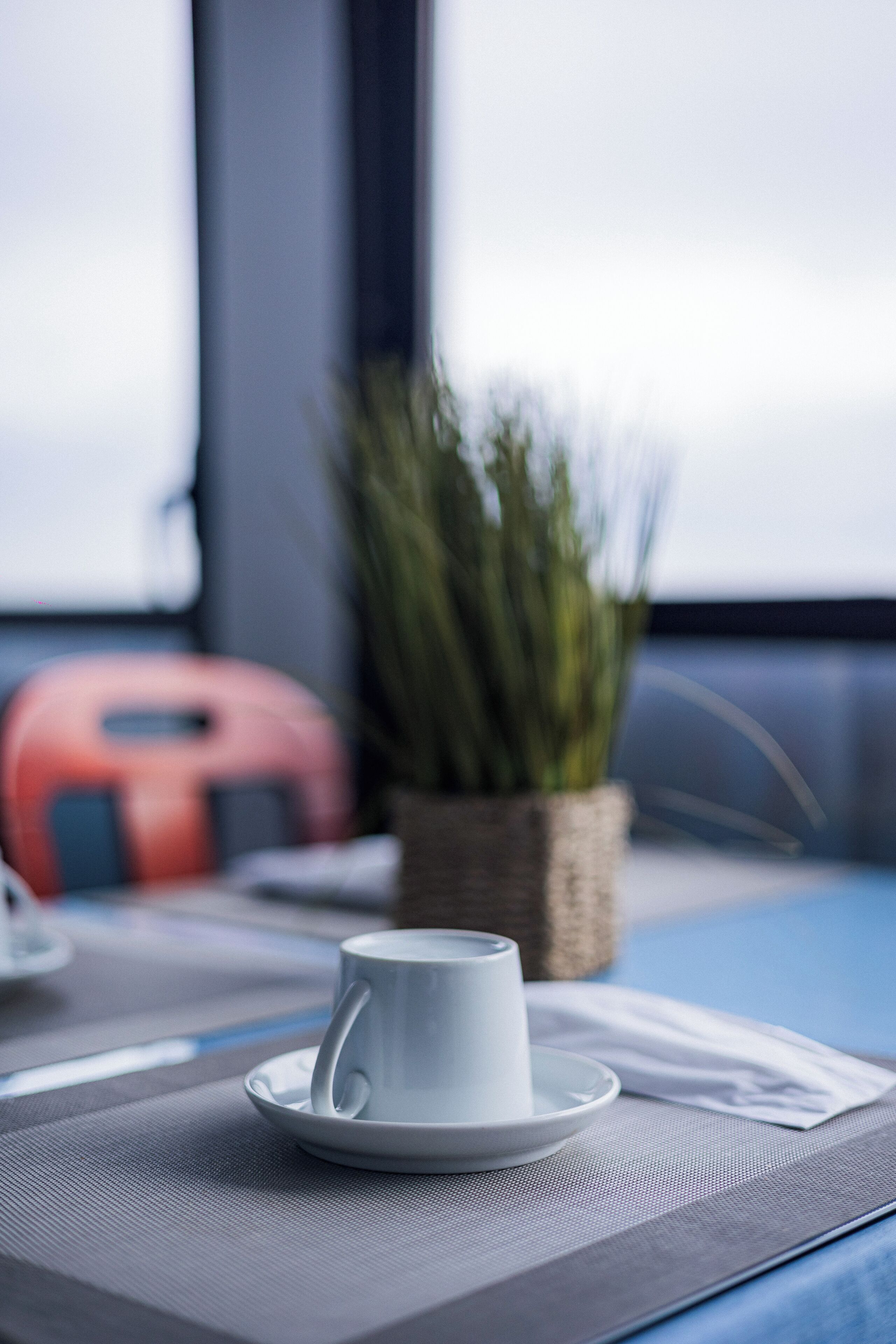 A white cup on a saucer on a gray placemat, a wicker basket with green plants, and a red object in the background.