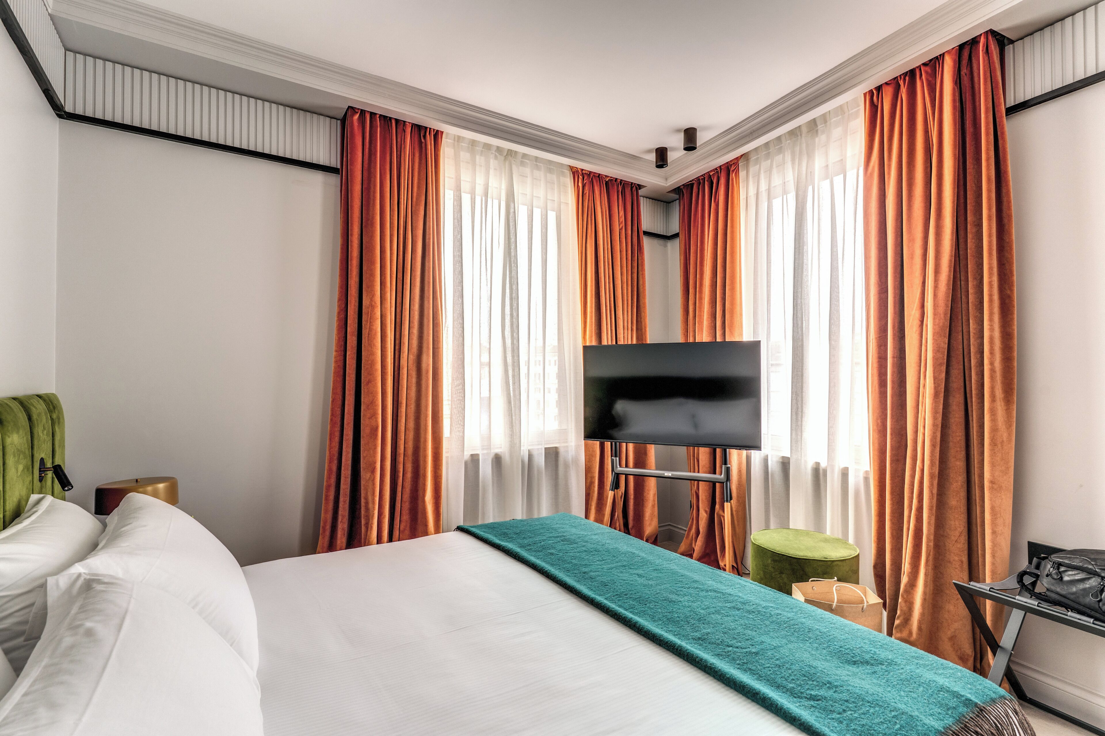 Camera d'albergo con un letto, una TV a schermo piatto, un poggiapiedi verde e un tavolino con una borsa.
