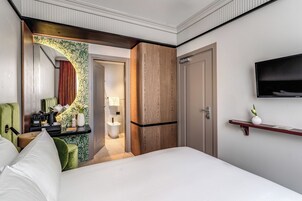 A hotel room with a bed, a desk with a lamp, a television, and a bathroom visible through an archway.