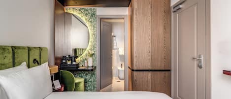 A hotel room with a bed, a green chair, a desk with a lamp, and a bathroom visible through an open door.