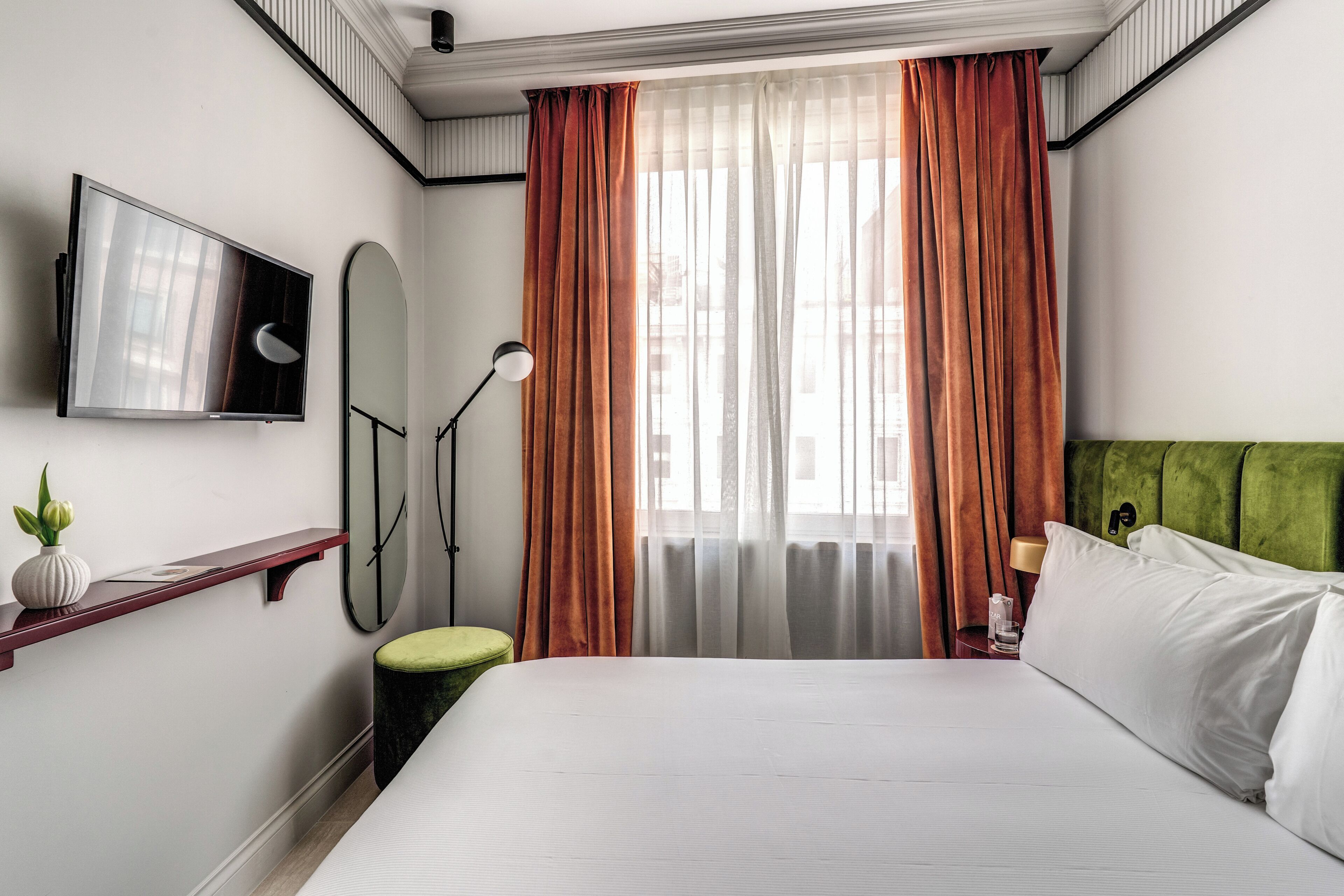 A hotel room with a bed, a TV, a desk, a lamp, and a window with curtains.