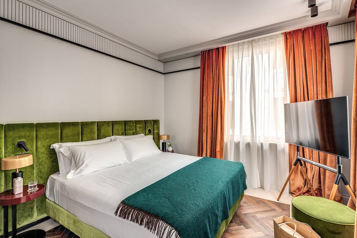 A hotel room with a green upholstered headboard, a bed with white linens, a teal throw blanket, a flat-screen TV on a stand, and a nightstand with a lamp.