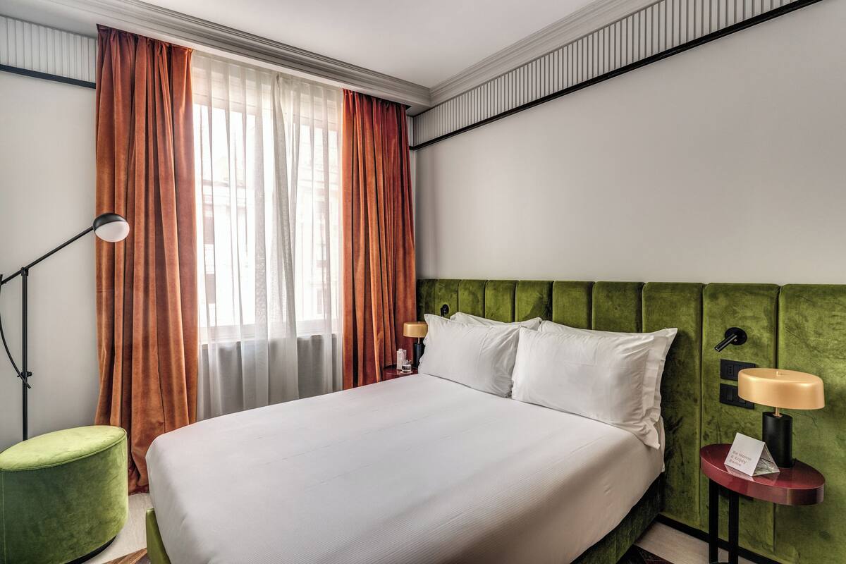 A hotel room with a large bed, green headboard, two bedside lamps, a green ottoman, and a window with curtains.