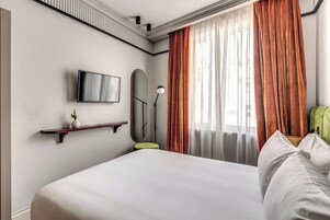 Et hotelværelse med en seng, et ophængt TV, et natbord med en lampe og et vindue med gardiner.
