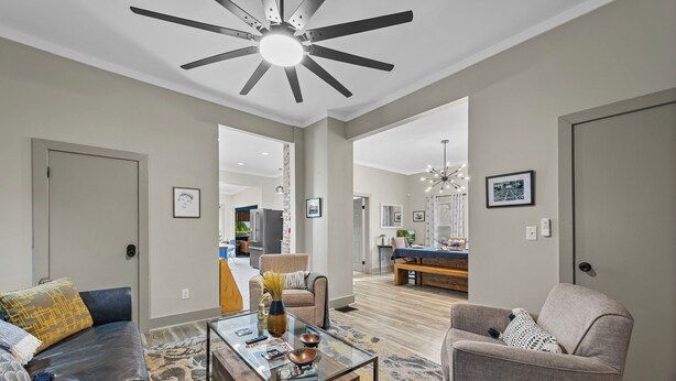 Ampla sala de estar com mesa de centro de vidro, sofá de couro e poltronas. É possível ver um ventilador de teto e um lustre.