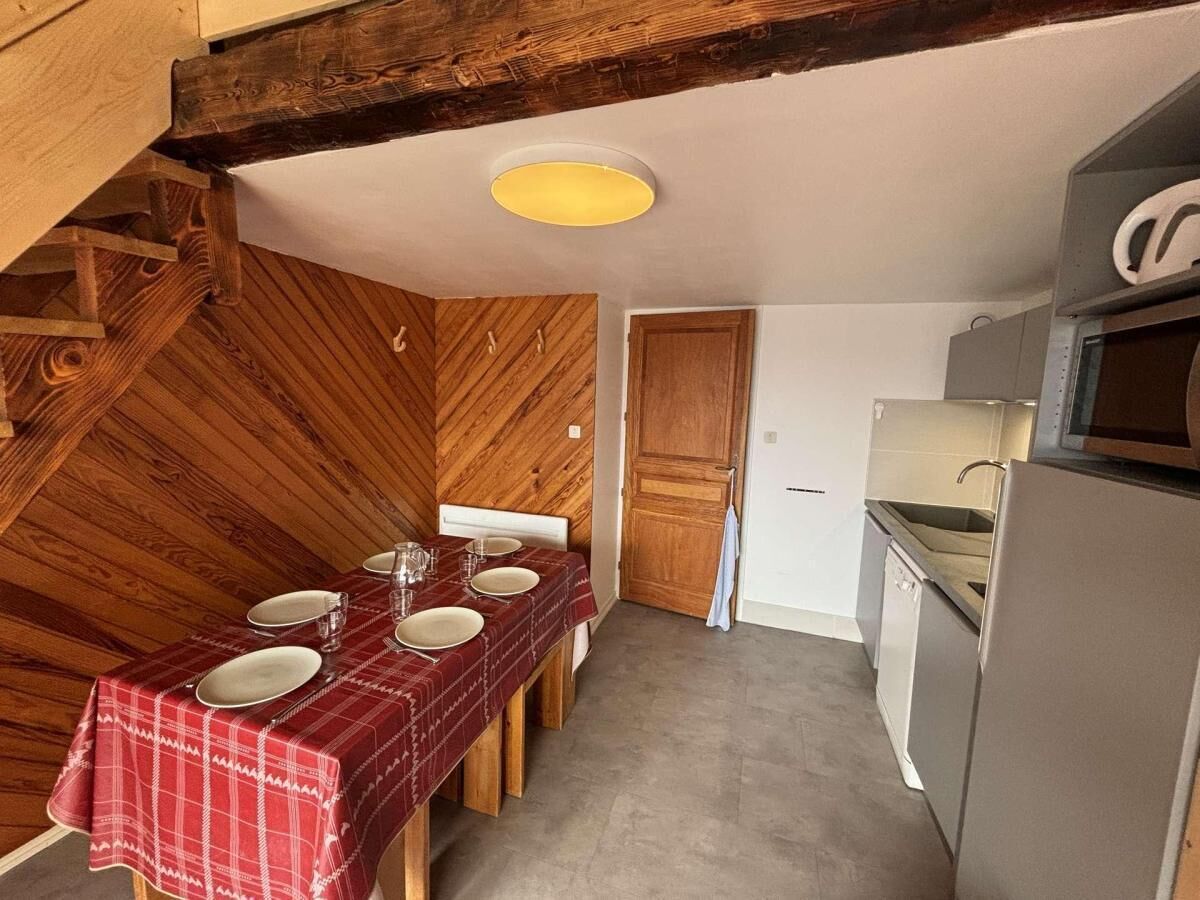 A dining table set with plates and glasses, a wooden wall, a kitchen with a refrigerator, and a door.