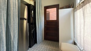 A compact kitchen with a refrigerator, oven, and a small sink. A door leads to an outdoor area with a brick wall.