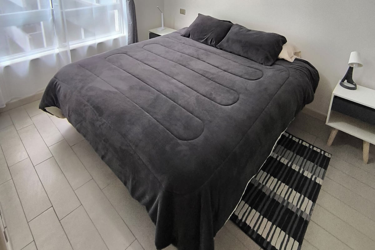 Une chambre moderne avec un grand lit, une table de chevet avec une lampe, une fenêtre avec des rideaux et une vue sur l’extérieur.