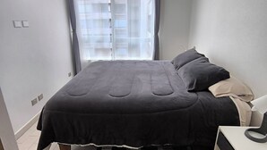 A bedroom with a bed, bedside table, lamp, and a window with curtains. - Audom Property Manager (Santiago)