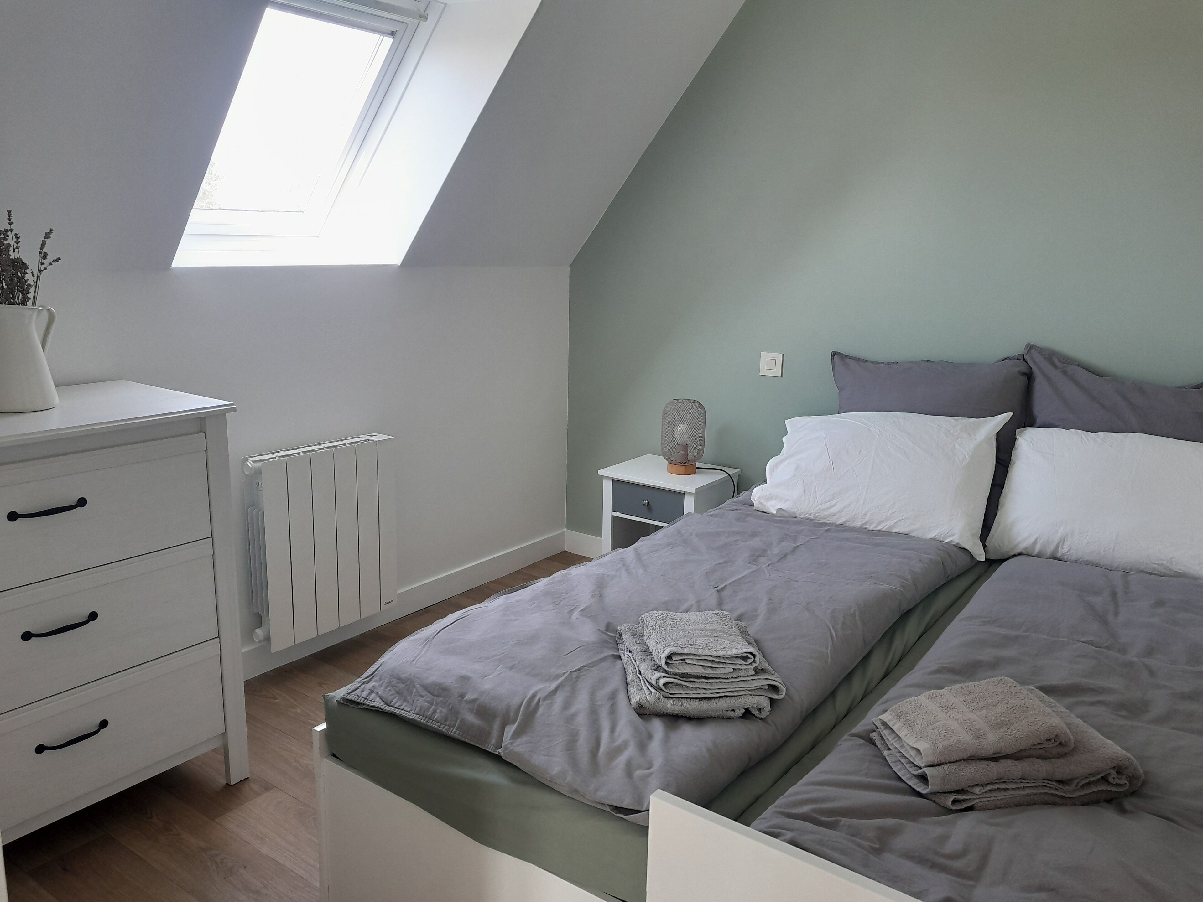 A bedroom with a sloped ceiling, a radiator, a bedside table with a fan, a bed with grey bedding and pillows, and a white dresser with drawers.