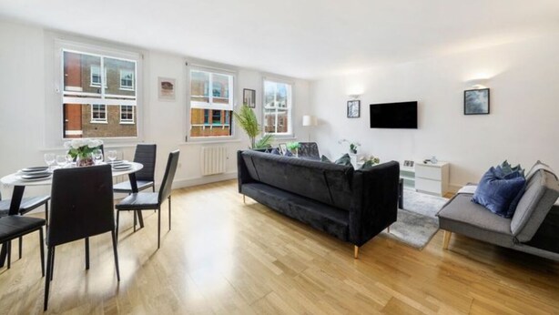 A modern living room with a dining area, large windows, a black sofa, and a flat-screen TV. - Trendy 2-Bed, 2-Bath flat near Soho, Marylebone & Oxford Circus (London)
