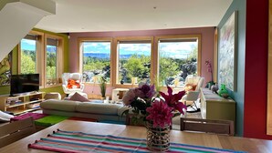 A spacious living room with a large window offering a mountain view, a cozy seating area with a sofa and chairs, and a dining table with a striped placemat.