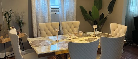 A dining area with a round table, white upholstered chairs, a chandelier, and a large plant.