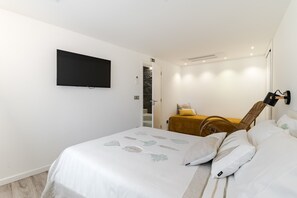 Ein Schlafzimmer mit einem Bett, einem flachbildfernseher an der Wand, einem Korbstuhl und einem kleinen Tisch mit einem gelben Kissen.