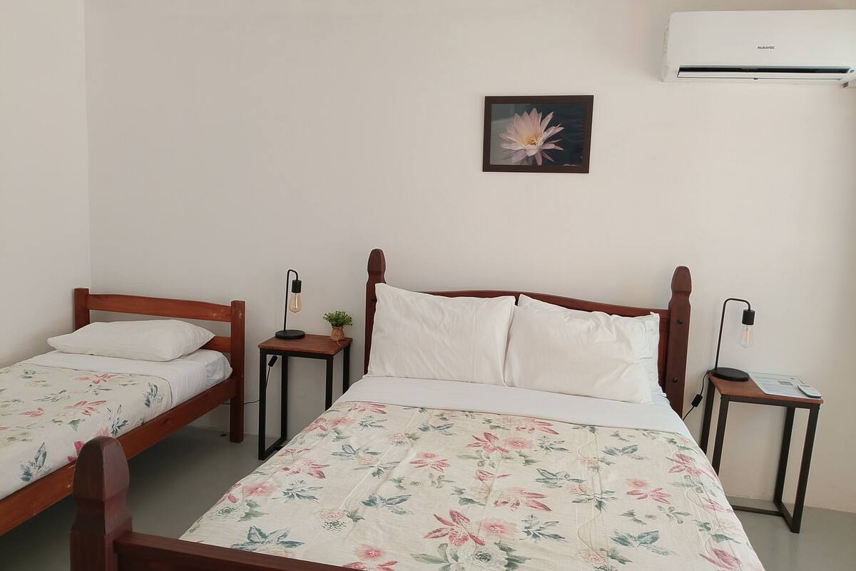 Quarto com duas camas de solteiro, mesa de cabeceira com abajur, pequena mesa com luminária e ar condicionado na parede.