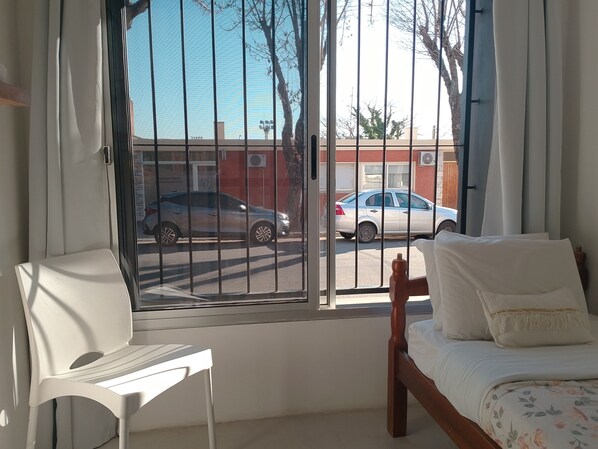 A bedroom with a bed, a chair, and a window with a view of parked cars and a building. - La casita de Tomi (Colonia del Sacramento)