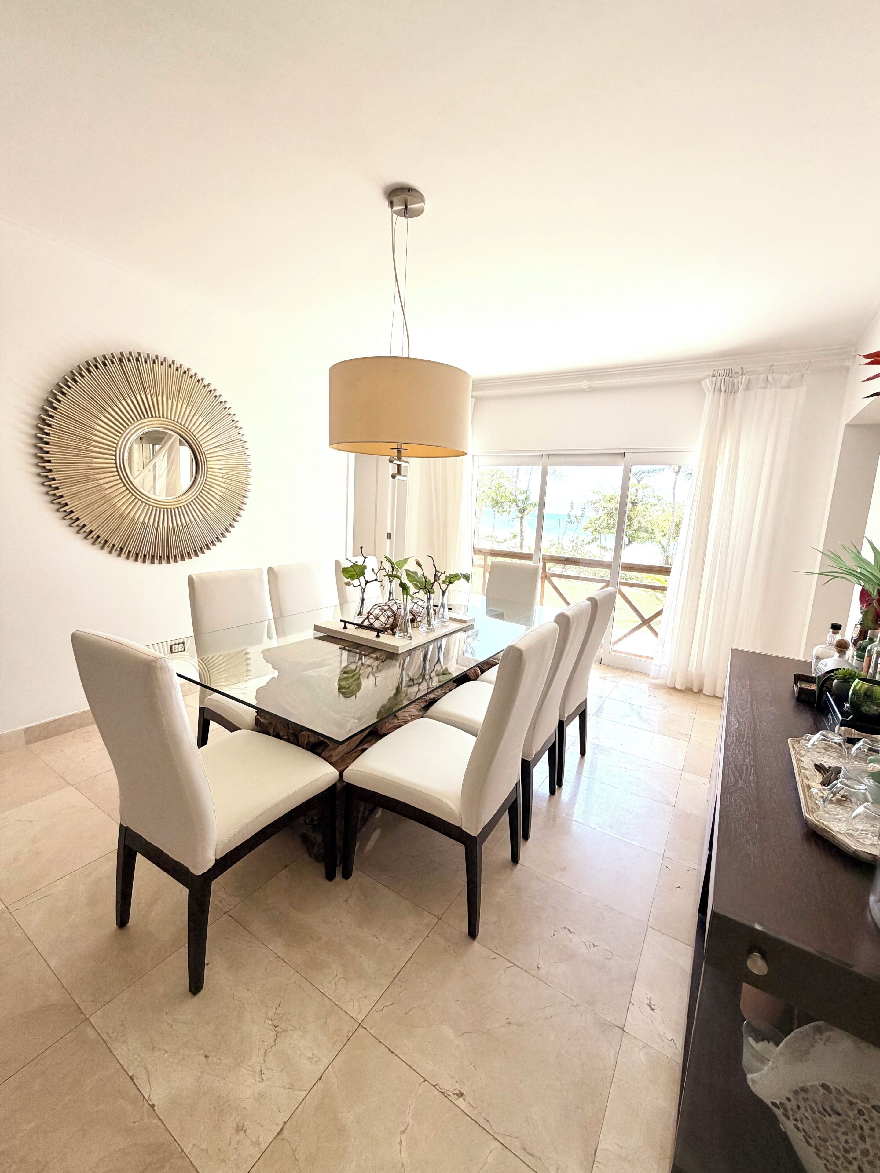Un comedor con mesa de vidrio, sillas blancas, una lámpara colgante y un espejo decorativo.