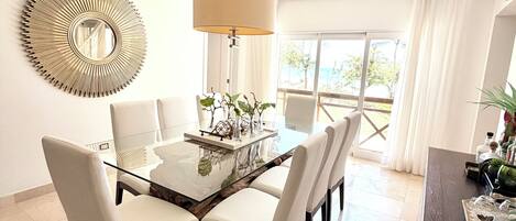 Un comedor con mesa de vidrio, sillas blancas, una lámpara colgante y un espejo decorativo.