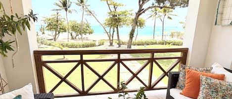 Un balcon donnant sur la plage, un canapé en osier avec des coussins, une table basse en bois agrémentée d’objets décoratifs et une rambarde.