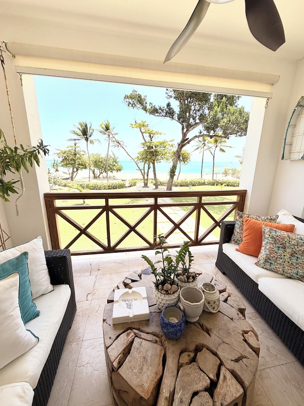 A balcony with a view of the beach, a wicker sofa with cushions, a wooden coffee table with decorative items, and a railing. - Riviera Azul - Playa Dorada (Puerto Plata)