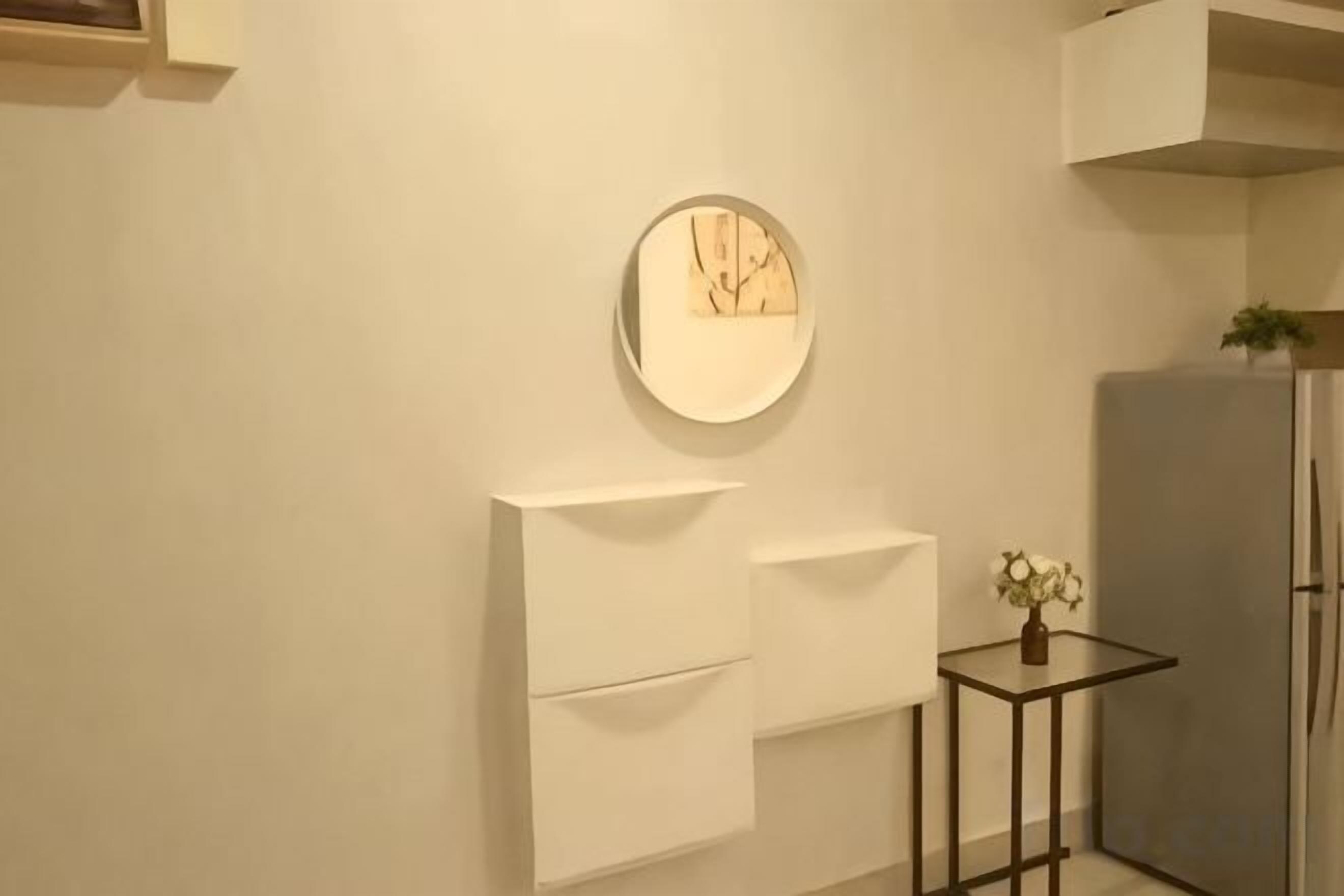 A kitchen with a white wall, a round mirror, a small table with a plant, and a refrigerator.