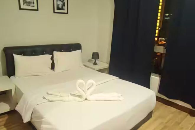 Una cama bien hecha con sábanas blancas y un arreglo de toallas con forma de cisne. Se observa una mesita de noche con una lámpara y una foto en un marco en la pared.