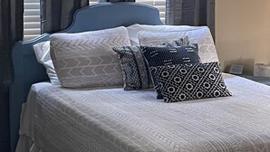 Een netjes opgemaakt bed met beddengoed en kussens, een blauw hoofdbord en een raam met gordijnen.