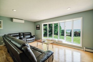 Amplia sala de estar con sofá de cuero, una mesa de centro y ventanas grandes que ofrecen vistas a un jardín y una masa de agua.
