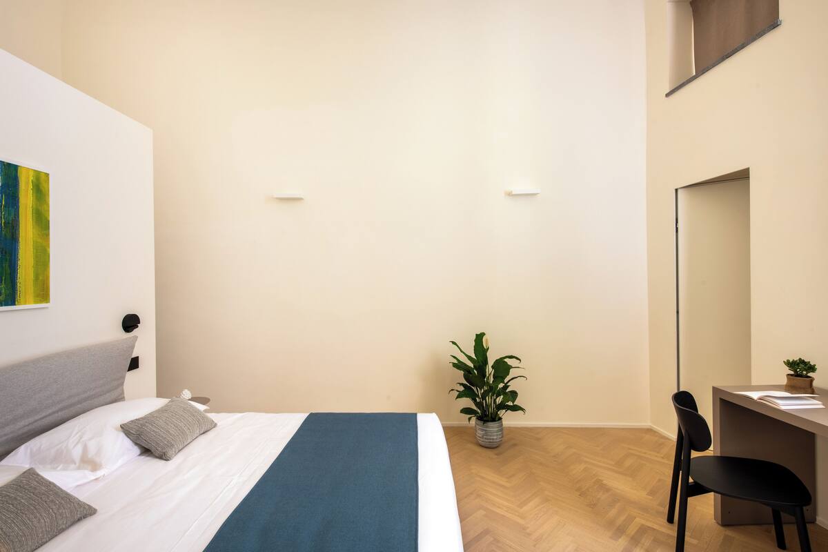 A modern hotel room with a bed, a desk, a chair, a potted plant, and a wall-mounted mirror.