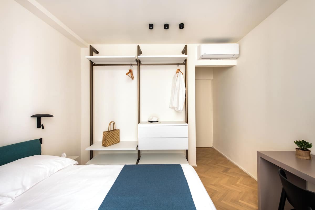 A hotel room with a bed, a desk, a hanging chair, a small table, a wall-mounted shelf, and an air conditioning unit.