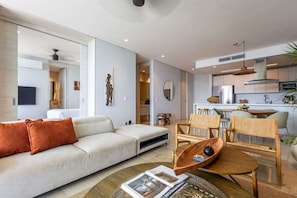 Un salon moderne avec un canapé, une table basse et des fauteuils. On aperçoit à l’arrière un coin cuisine avec des placards et des appareils électroménagers.