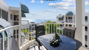 A balcony with a table, chairs, and binoculars, overlooking a coastal area with buildings and the sea.