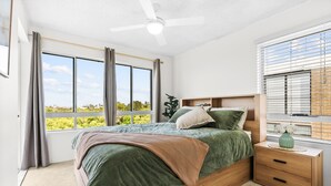 A bedroom with a large bed, a bedside table, a nightstand, a ceiling fan, and a window with curtains.