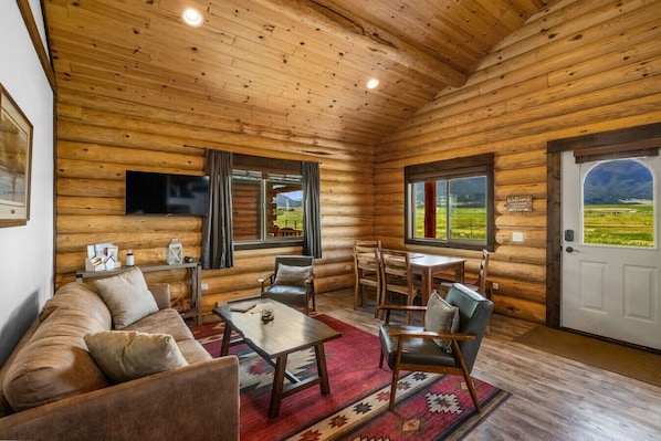 L’intérieur d’une cabane en rondins, avec un canapé, des fauteuils, une table, un téléviseur et une porte donnant sur un champ.