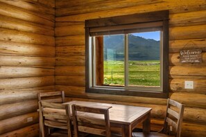 Une cabane en bois avec une table et des chaises, une fenêtre donnant sur un paysage verdoyant et des montagnes, et une pancarte de bienvenue.