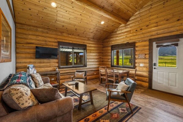A log cabin interior with a sofa, chairs, a table, a TV, and windows with a view.