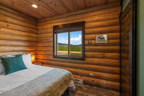 Une chambre à coucher dans une cabane en rondins, avec un lit, une fenêtre donnant sur l’extérieur et un tableau encadré représentant un élan accroché au mur.