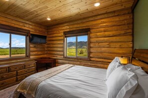 A log cabin bedroom with a bed, a desk, a TV, and windows with a mountain view. - Mountain View Cabin in Pray MT (Pray)