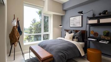 A modern bedroom with a large window, a bed with grey bedding, a bench, a desk with a lamp, and a shelf with decorative items.
