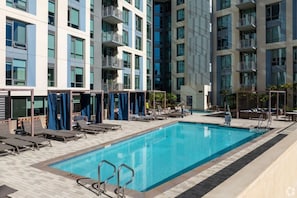 Un espace piscine aménagé sur le toit, avec des chaises longues et une piscine, le tout entouré d’un immeuble moderne doté de balcons.