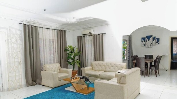A living room with two beige sofas, a coffee table, and a blue rug. There is a dining area with a table and chairs in the background. - VIST Two-Bedroom Apartment with Pool View unit 1 (Accra)