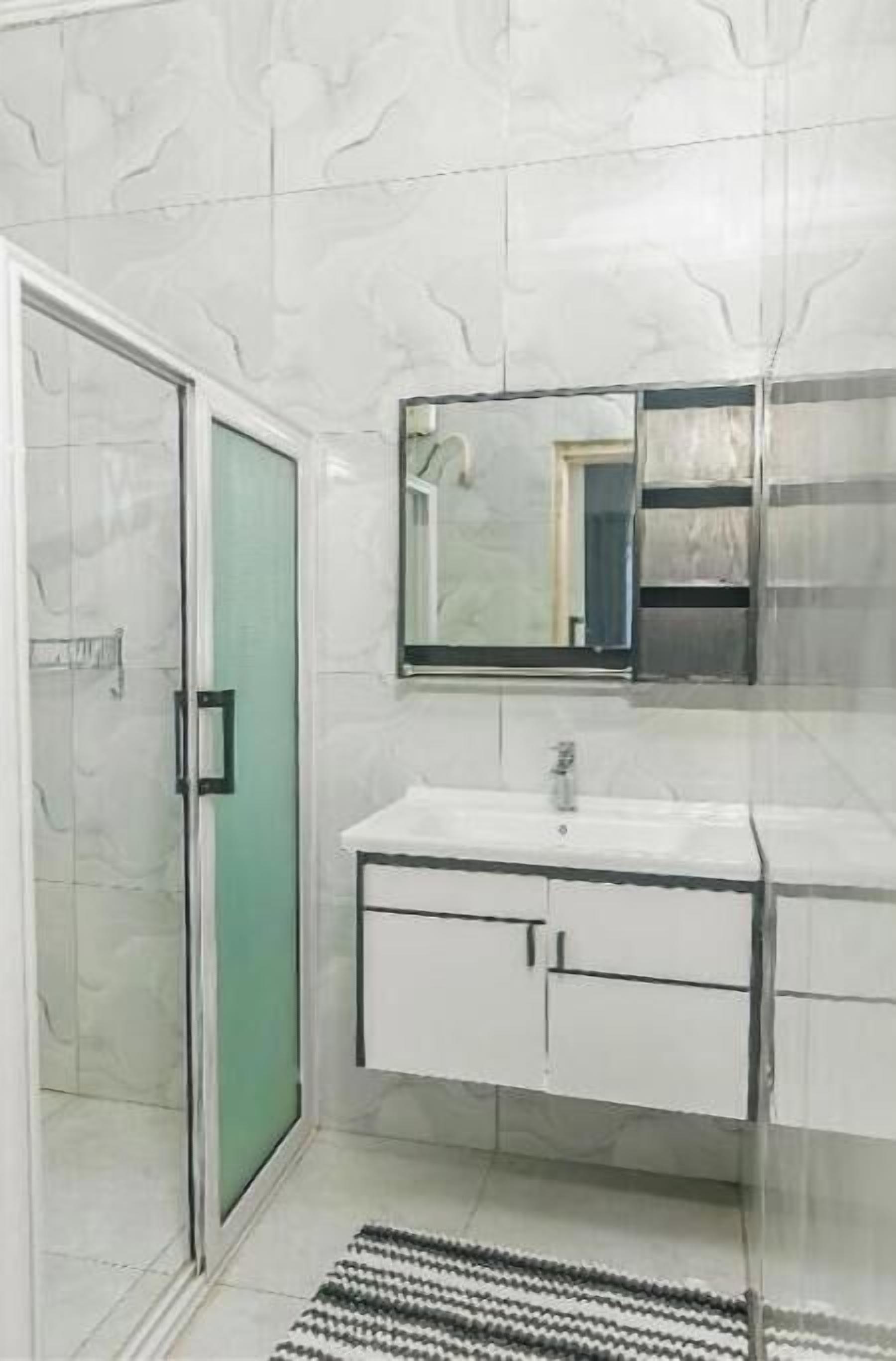 Un baño con mampara de vidrio, un lavamanos blanco con un espejo encima y piso de baldosas.