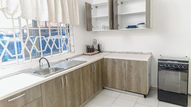 A kitchen with wooden cabinets, a sink, a stove, and a window with a view of a blue sky and a white fence. - VIST One-Bedroom Apartment with Pool View (Accra)