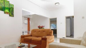 A living room with a brown sofa, a glass coffee table, and a beige armchair. There is a painting on the wall and a vase with flowers on a table. - VIST One-Bedroom Apartment with Pool View (Accra)