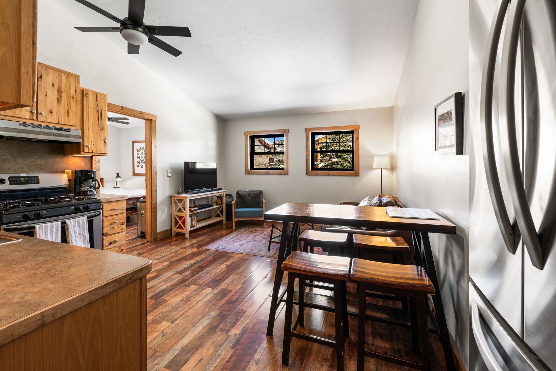 A compact kitchen with wooden cabinets, a stove, and a ceiling fan. There is a dining area with a table and chairs, and two windows with artwork.