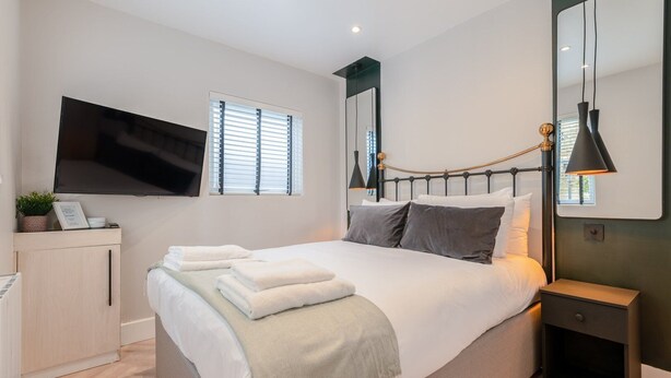 A modern bedroom with a large bed, bedside tables, a TV mounted on the wall, and a window with blinds. - The Balham Hideaway (London)