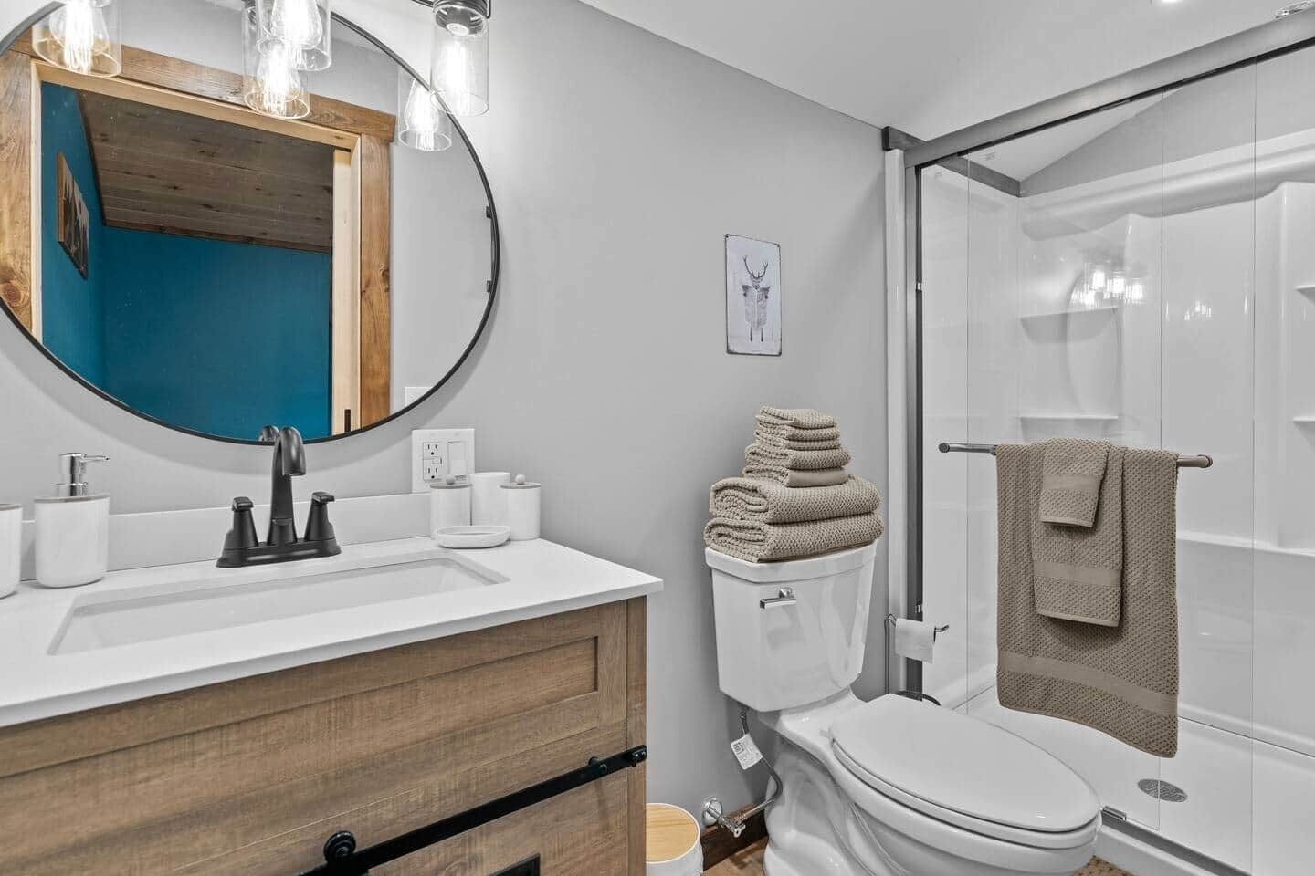 A bathroom with a wooden vanity, a round mirror, a toilet, and a glass shower enclosure.