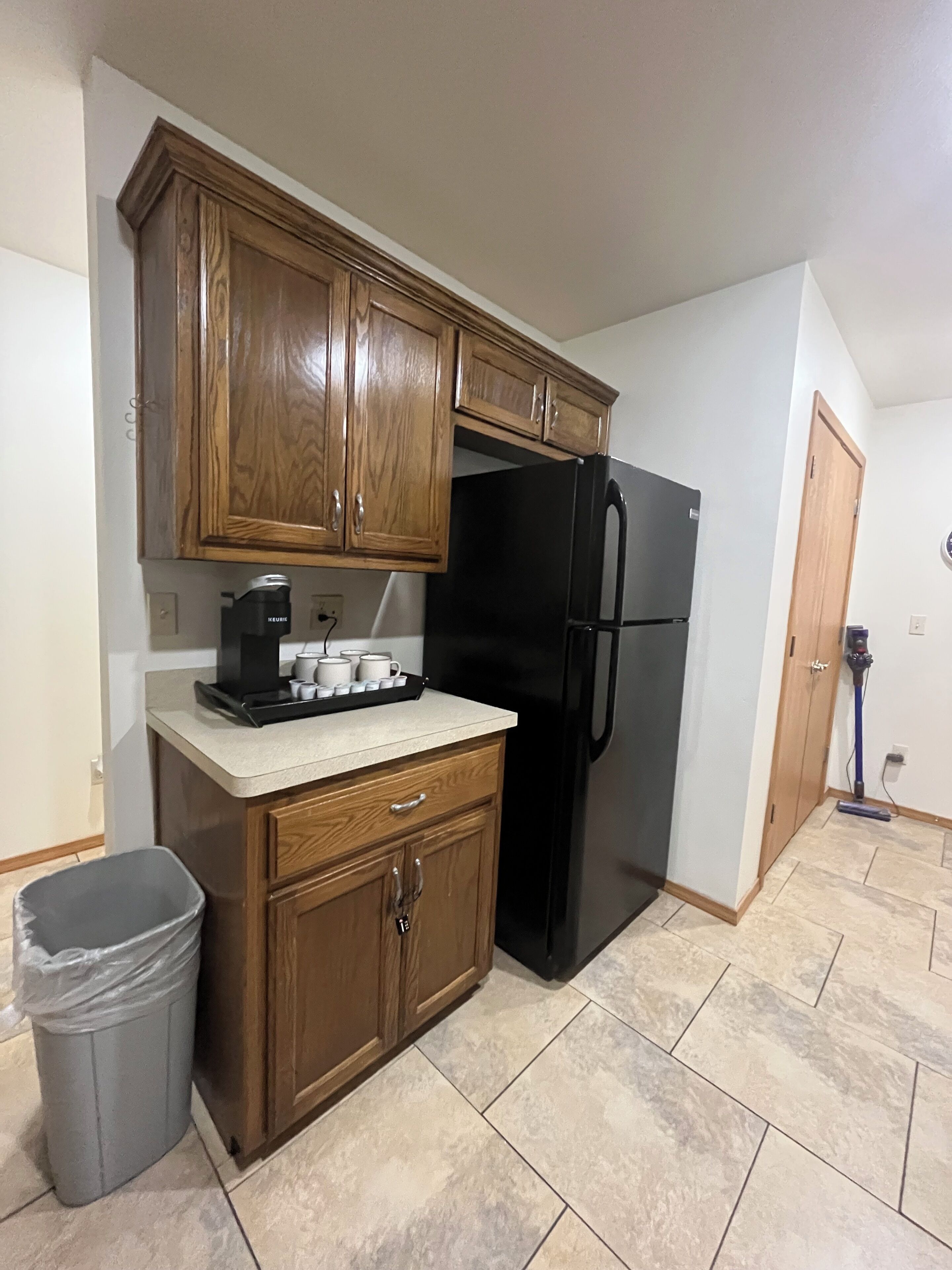 A compact kitchen with wooden cabinets, a black refrigerator, a coffee maker, and a trash bin.