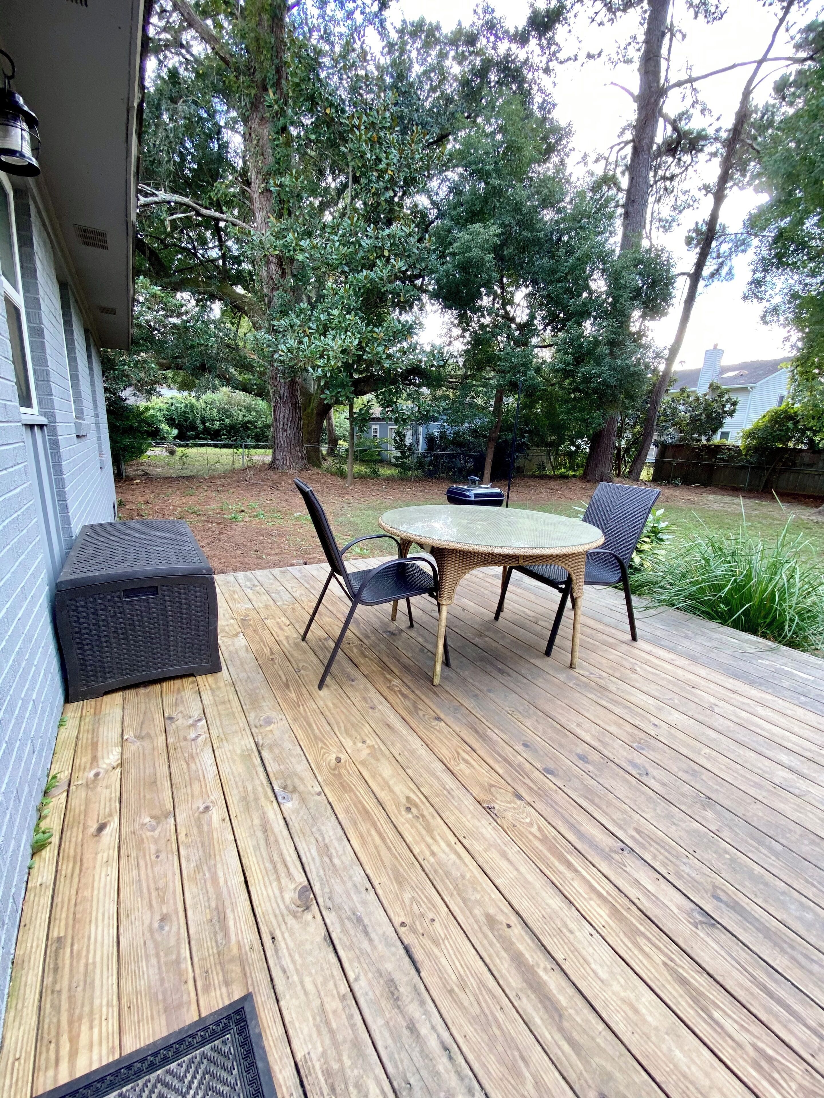 Una terraza de madera con una mesa y sillas, rodeada de árboles y una casa.