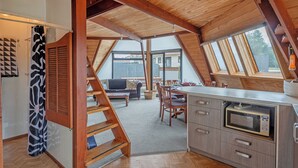 A cozy attic bedroom with a wooden ceiling, a built-in desk with a microwave, and a view of the backyard through large windows.